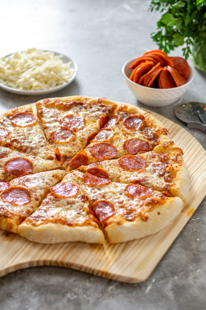 A homemade pizza sitting on a wooden pizza peel at an angle showing off the melted cheese.