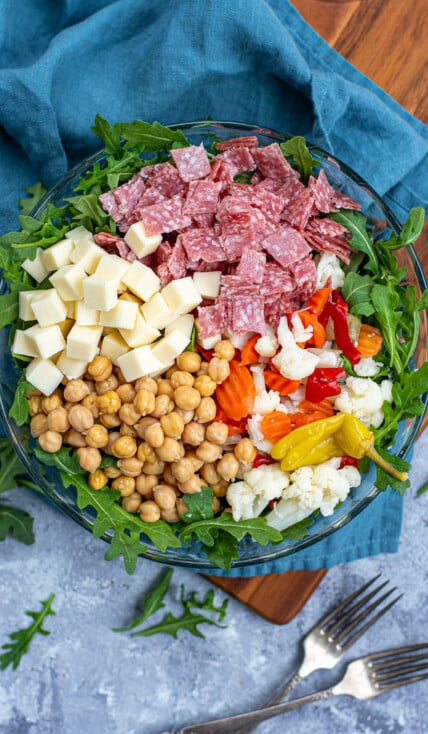 Overhead view of a glass bowl with the toppings for easy antipasto salad laid out evenly over a bed of arugula lettuce.