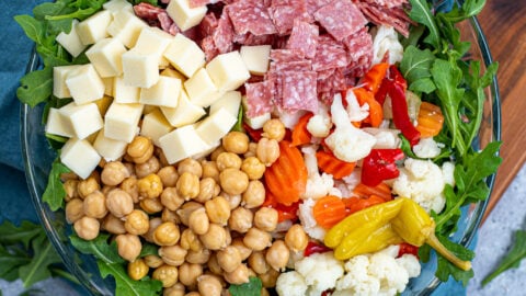 Overhead view of a glass bowl with the toppings for easy antipasto salad laid out evenly over a bed of arugula lettuce.