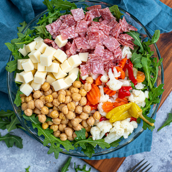 Overhead view of a glass bowl with the toppings for easy antipasto salad laid out evenly over a bed of arugula lettuce.