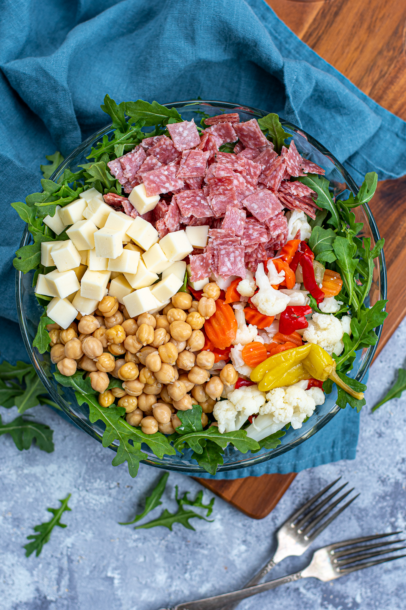 Overhead view of a glass bowl with the toppings for easy antipasto salad laid out evenly over a bed of arugula lettuce.