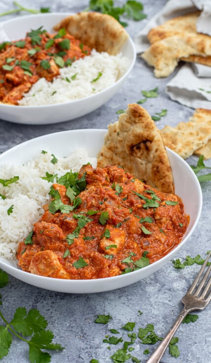 Chicken Tikka Masala is a large white bowl next to a scoop of rice and with a torn piece of naan.