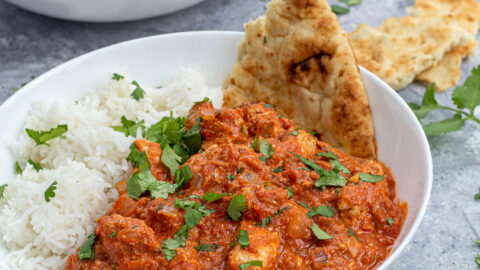 Chicken Tikka Masala is a large white bowl next to a scoop of rice and with a torn piece of naan.