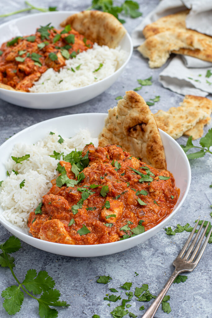 Chicken Tikka Masala is a large white bowl next to a scoop of rice and with a torn piece of naan.