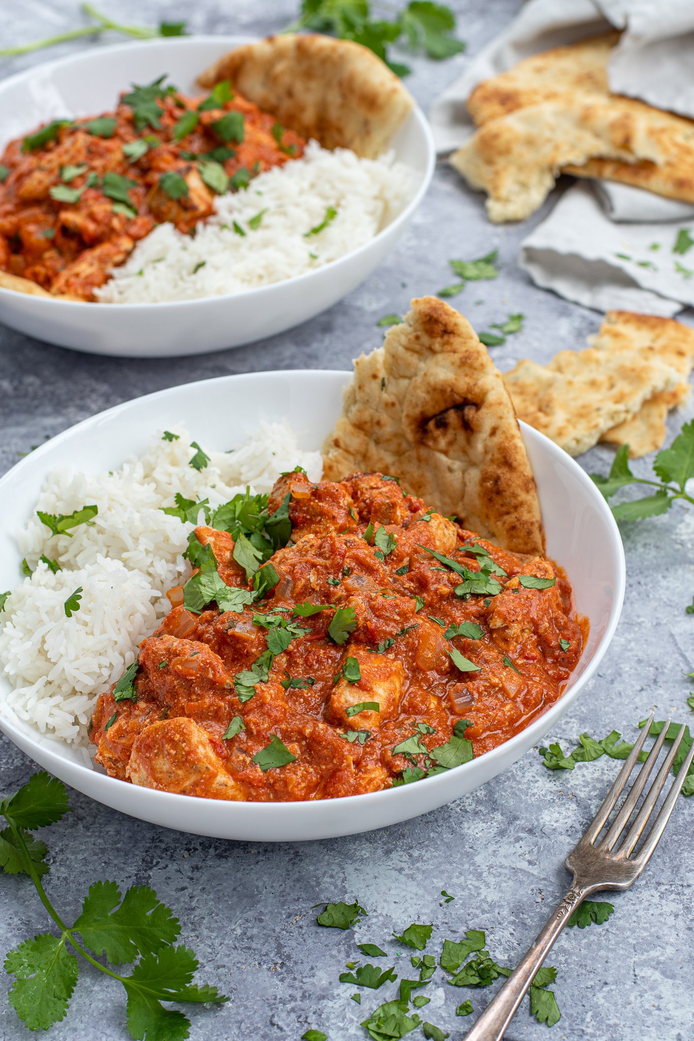 Chicken Tikka Masala is a large white bowl next to a scoop of rice and with a torn piece of naan.