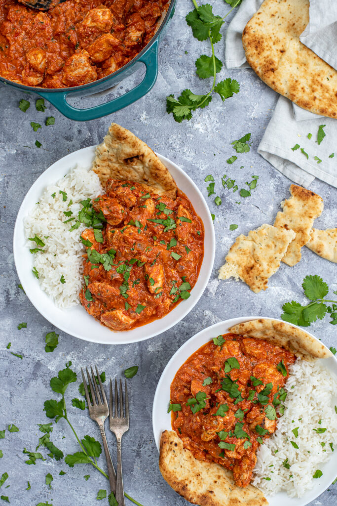 Flatly of two bowls and a dutch oven full of bright chicken tikka masala, covered with sprinkles of cilantro and pieces of naan bread.