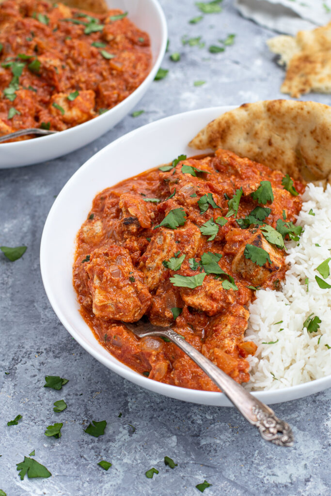 A fork in a large white bowl of chicken tikka masala next to a large scoop of rice and sprinkled with cilantro.