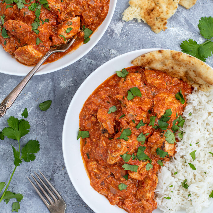 Overhead close up of chicken tikka masala in a large white bowl next to a bed of rice covered with sprinkles of cilantro.