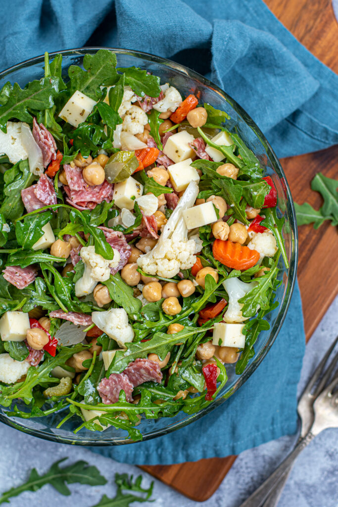 Overhead view of a tossed antipasto salad in a glass bowl showing off salami, cheese, chickpeas, and pickled Italian vegetables.