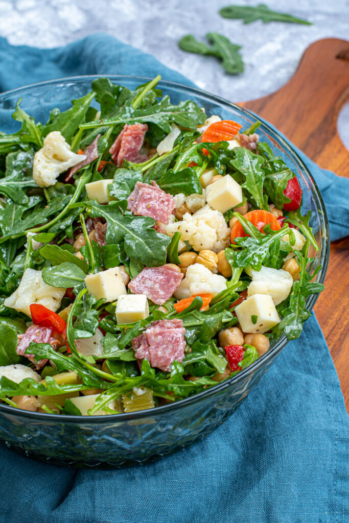 A tossed easy antipasto salad in a glass salad bowl showing off cheese cubes, salami, and pickled vegetables.