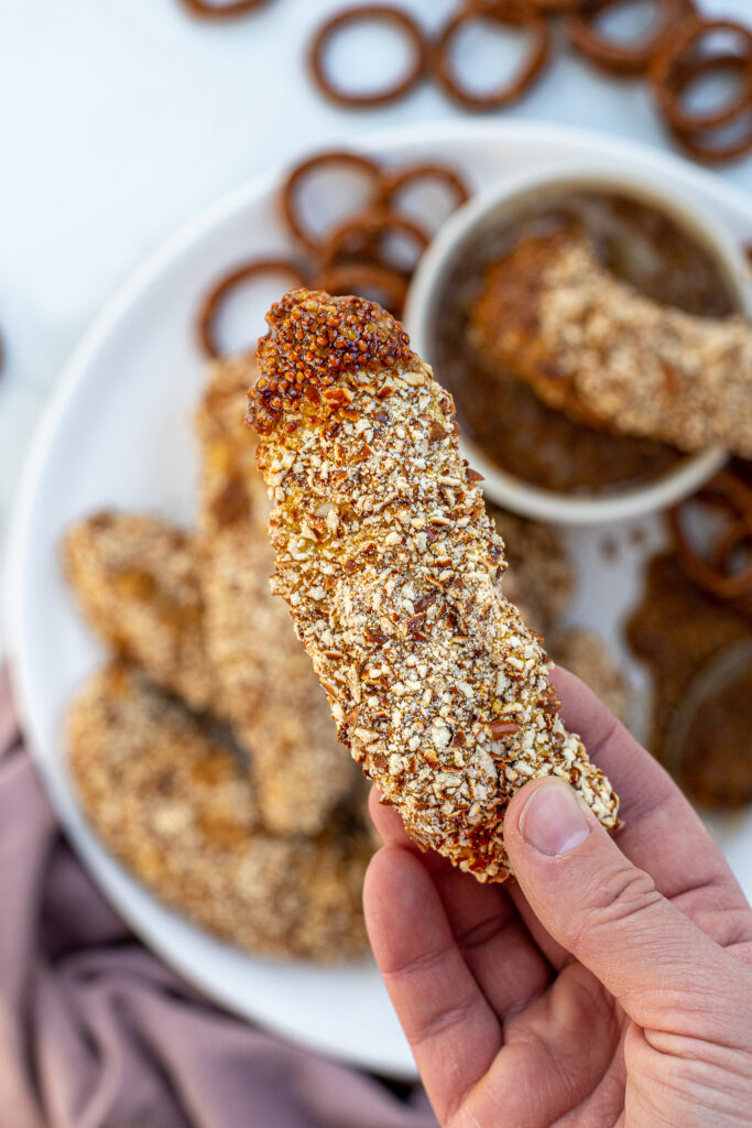 Hand holding a pretzel coated chicken tender that way dipped in honey mustard sauce.