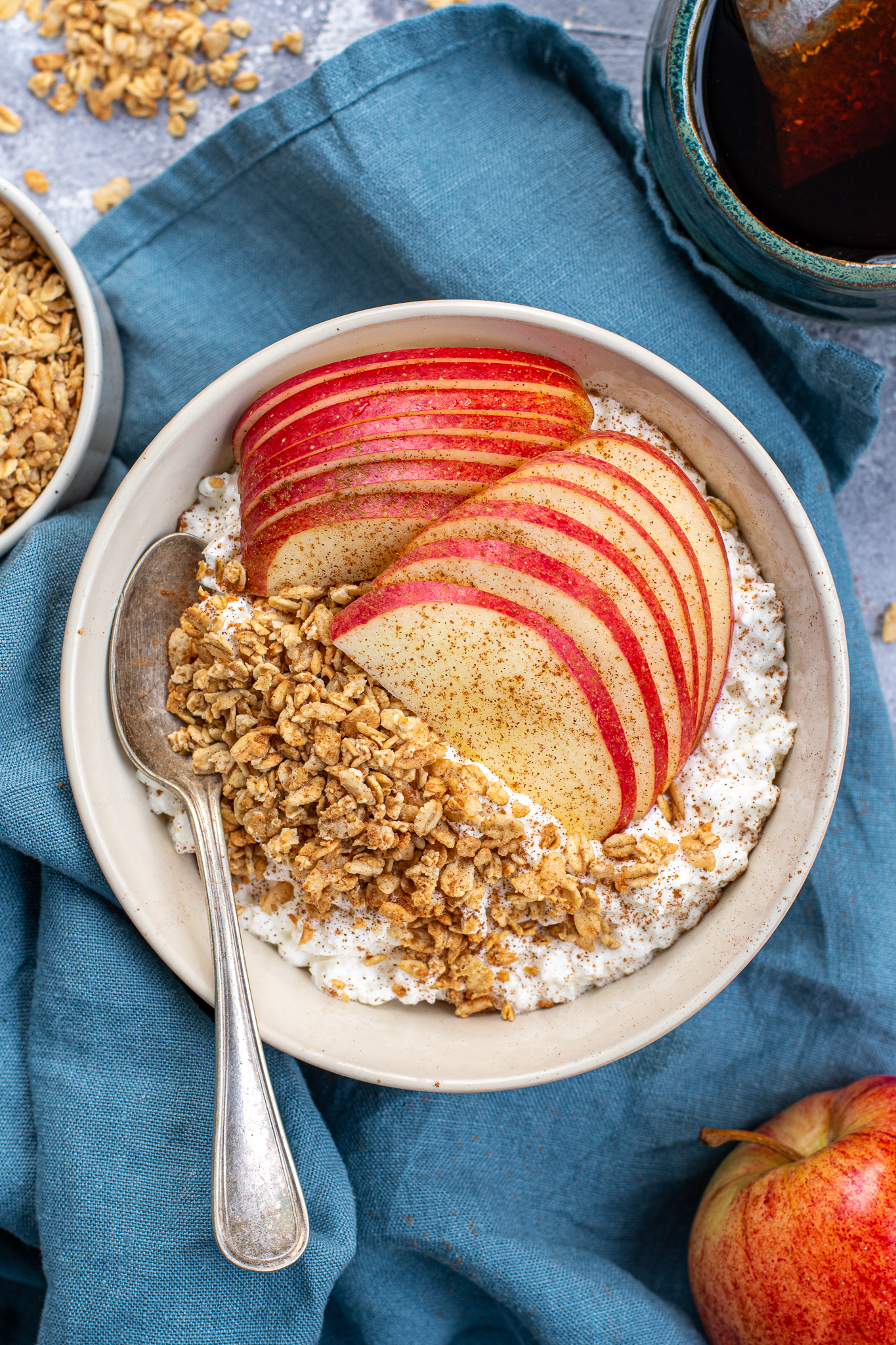 A white bowl on a blue towel filled with cottage cheese and apples with granola and ground cinnamon with a spoon ready to eat.