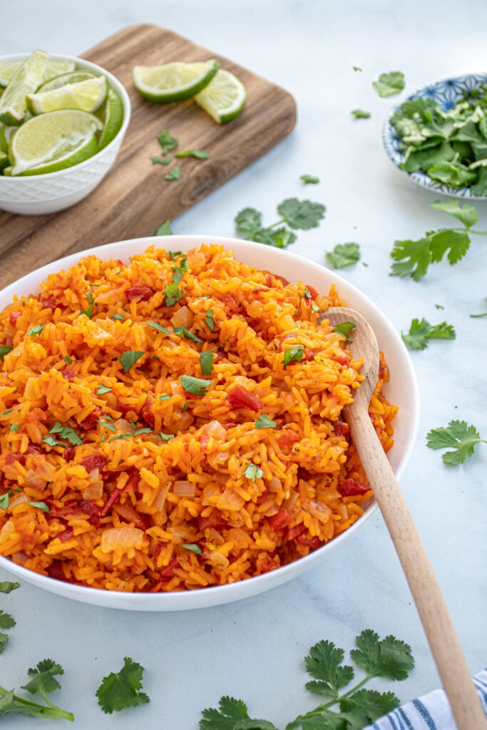 Large white bowl filled with red-orange colored mexican rice garnished with cilantro with a wooden spoon.