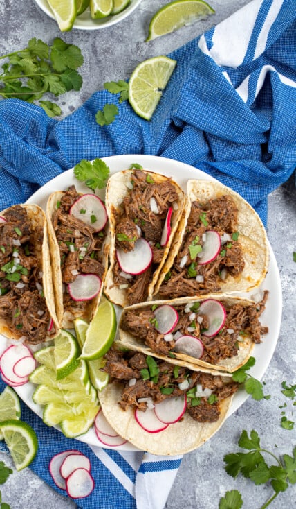 A tabletop with a blue towel, limes, cilantro, and a white plate with a bunch of tacos that are filled with crockpot beef barbacoa.