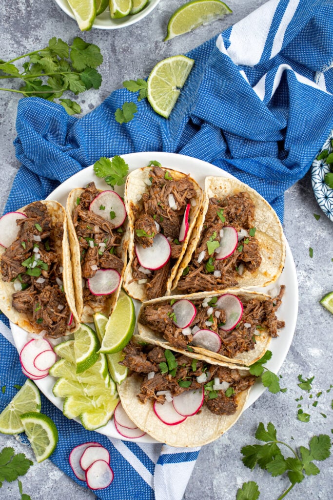 A tabletop with a blue towel, limes, cilantro, and a white plate with a bunch of tacos that are filled with crockpot beef barbacoa.