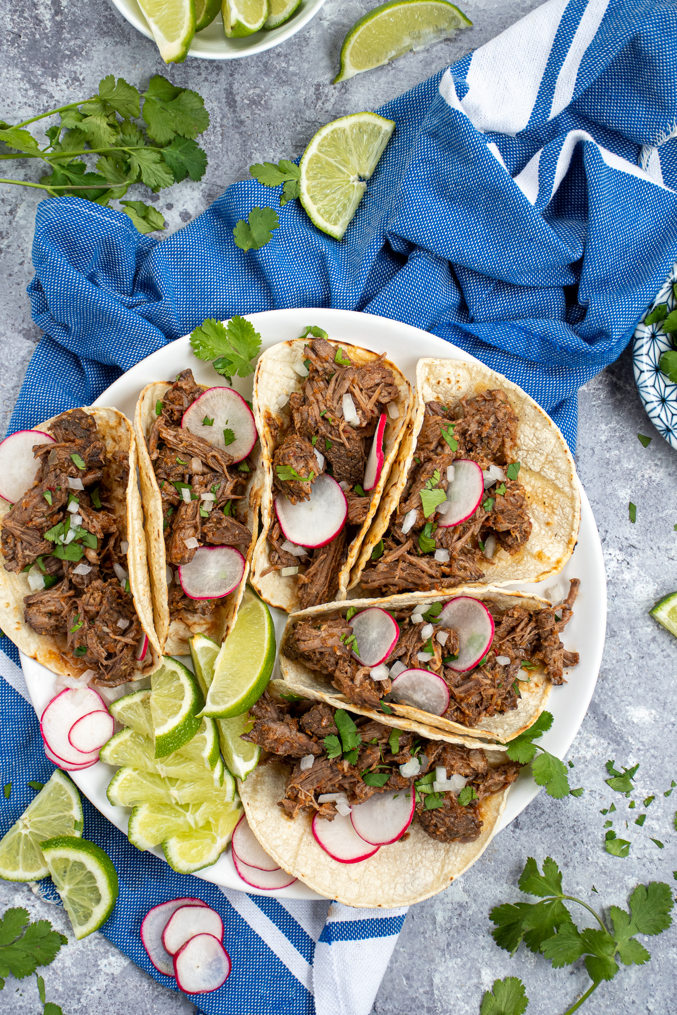 A tabletop with a blue towel, limes, cilantro, and a white plate with a bunch of tacos that are filled with crockpot beef barbacoa.
