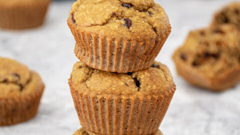 A stack of 3 blender flourless banana muffins with mini chocolate chips making a muffin tower with a plate of muffins in the background.
