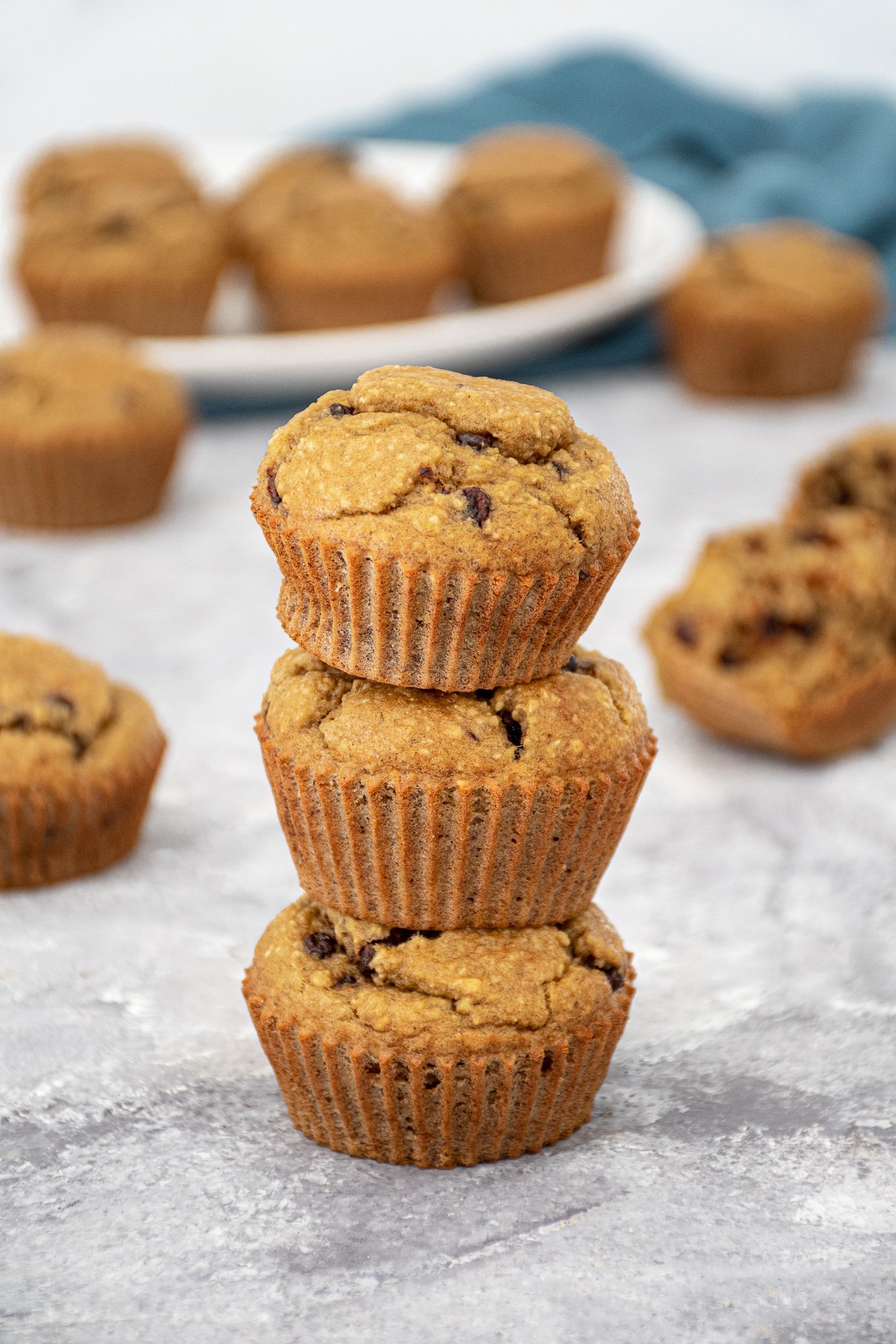 A stack of 3 blender flourless banana muffins with mini chocolate chips making a muffin tower with a plate of muffins in the background.