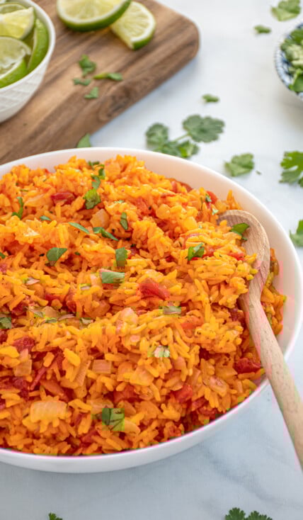 Bright orange-red colored Mexican Rice in a large white bowl with a wooden spoon garnished with chopped cilantro.