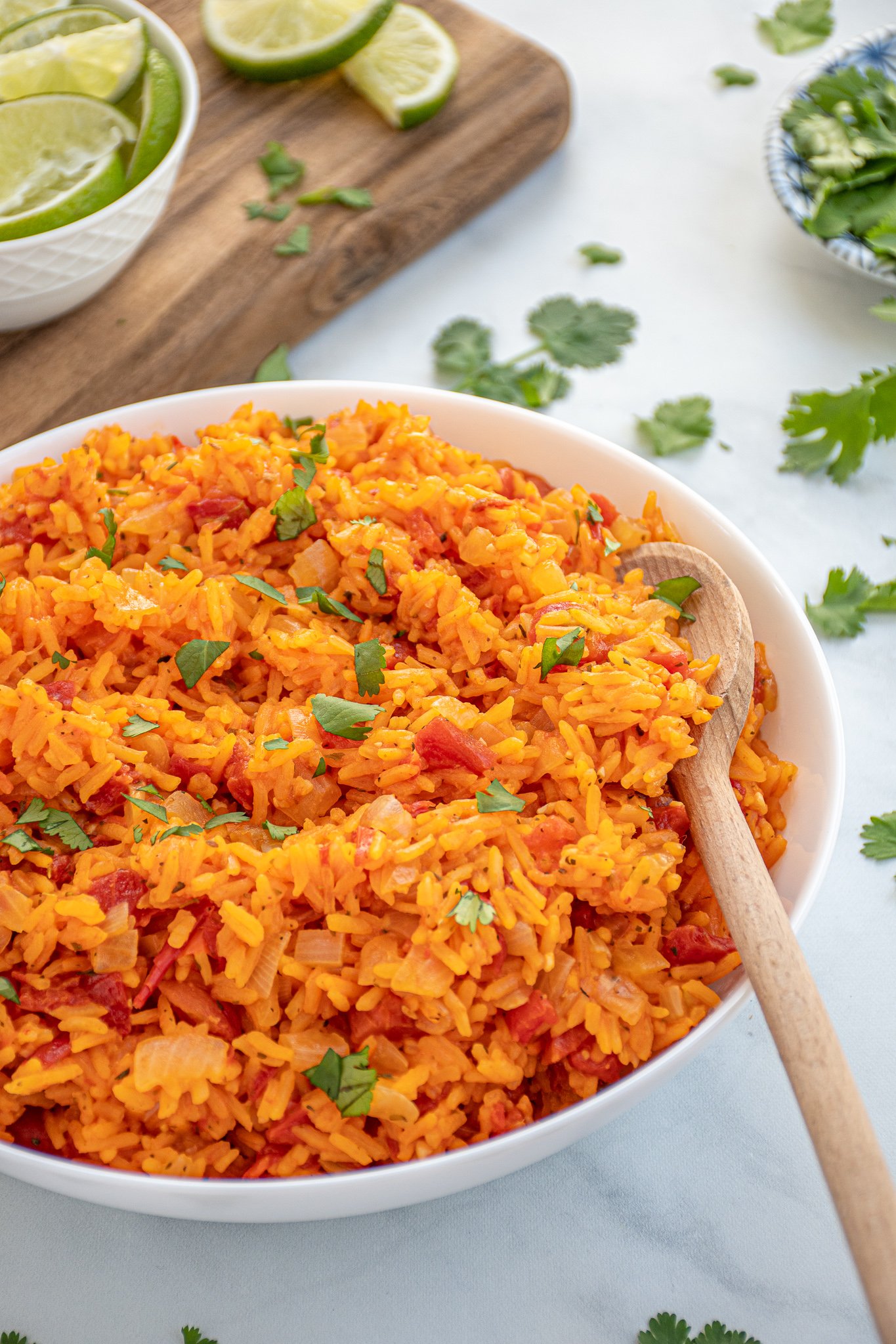 Bright orange-red colored Mexican Rice in a large white bowl with a wooden spoon garnished with chopped cilantro.