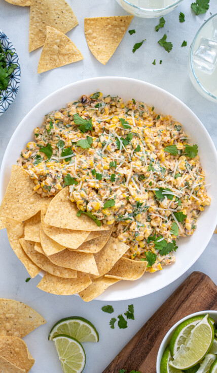 Large what bowl filled with Mexican Street Corn Dip and tortilla chips surrounded by more chips, cilantro, and limes.