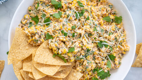Large what bowl filled with Mexican Street Corn Dip and tortilla chips surrounded by more chips, cilantro, and limes.