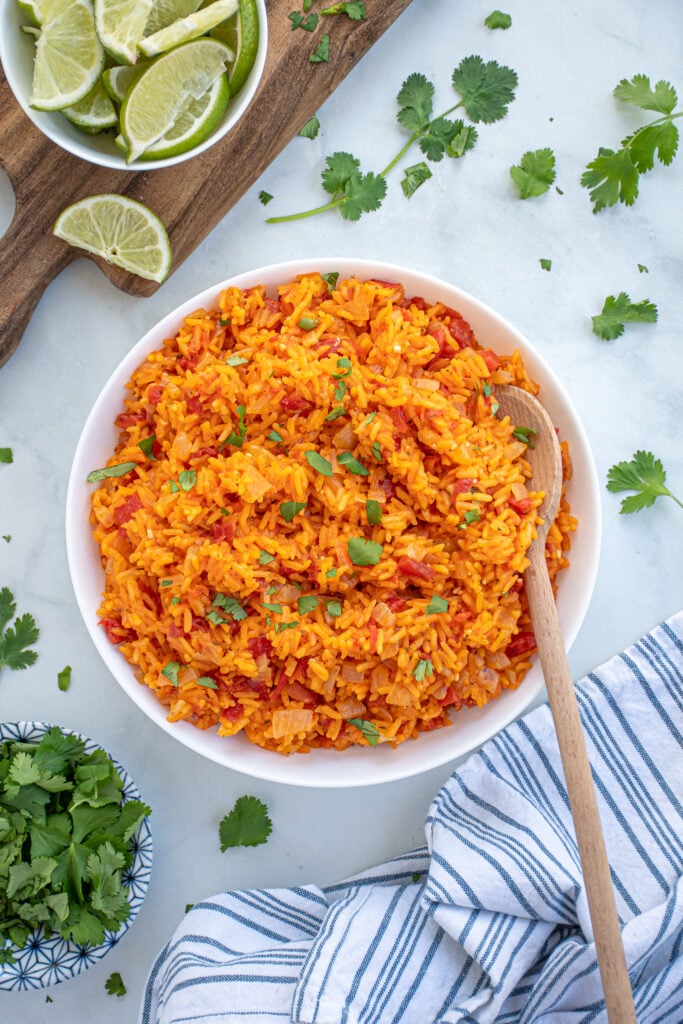 Overhead view of a large white bowl of mexican rice on a table surrounded by cilantro and cut up limes.