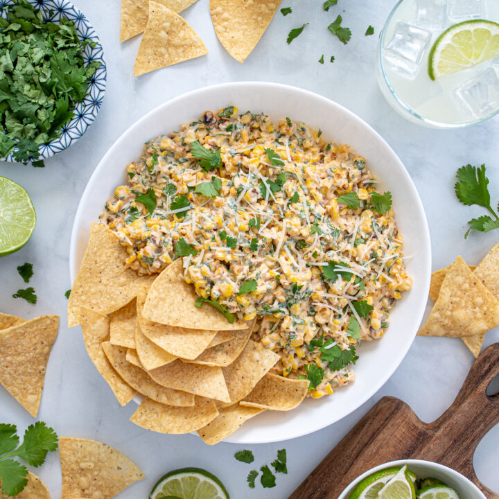 Overhead view of a table scene with a large bowl of Mexican street corn dip with tortilla chips surrounded by cut limes, cilantro pieces, and margaritas.