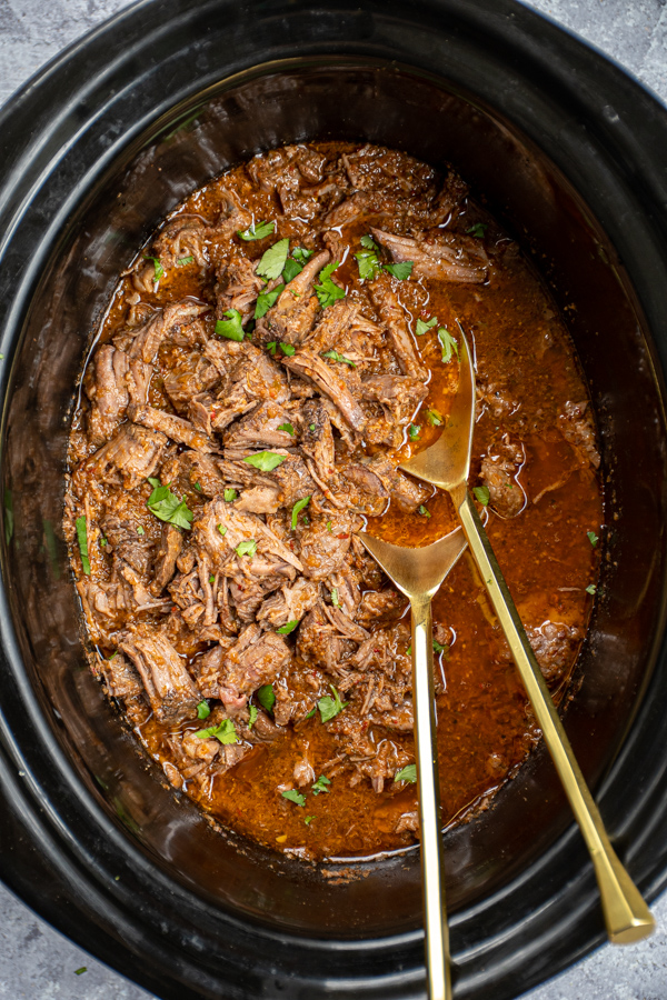 Close look inside a black crockpot showing shredded beef barbacoa garnished with cilantro and two golden spoons for serving.