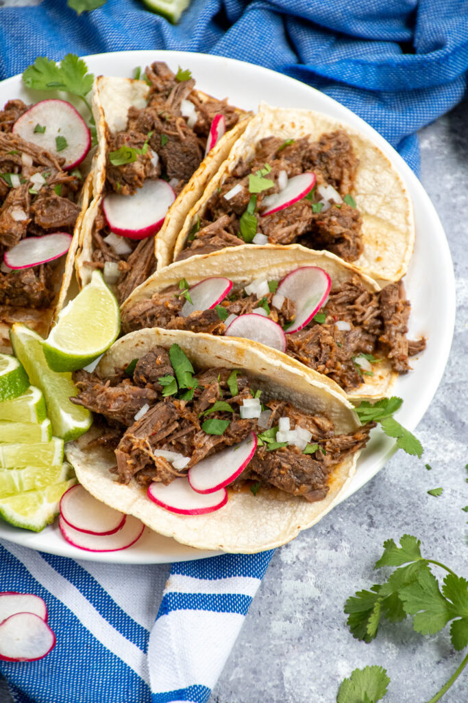 Side angle of a white plate filled with a bunch of tacos filled with crockpot barbacoa and garnished with onions, radishes, and cilantro.