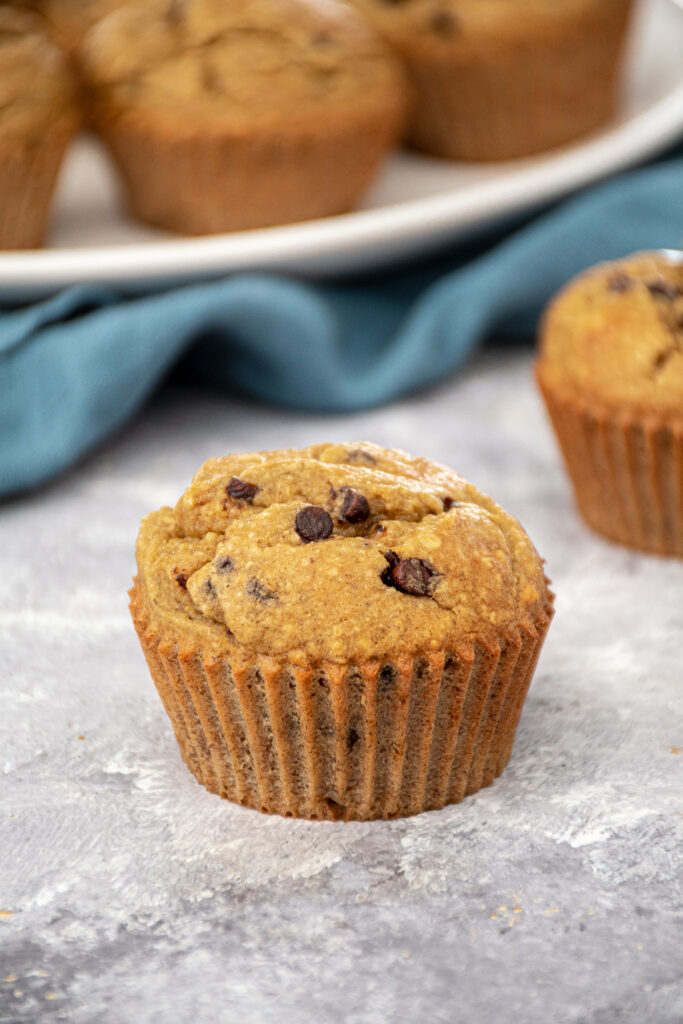 A single Flourless Banana Blender Muffin with mini chocolate chips sitting on a countertop with a plate of muffins sitting in the background.