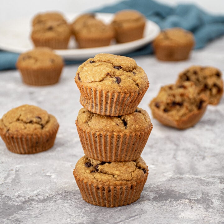 Three flourless banana muffins with chocolate chips stacked on one of another to make a muffins tower with a plate of muffins in the background.