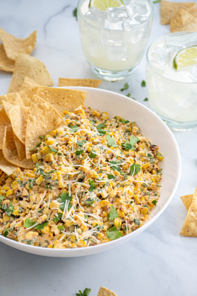 Angled view of a table with a bowl of street corn, tortilla chips, and margaritas.