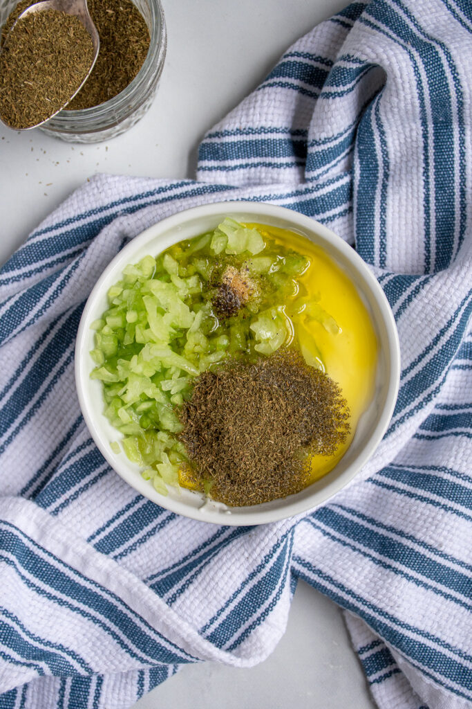 Small bowl on a blue kitchen towel sit the ingredients for tzatziki all placed in the bowl so you can see them.