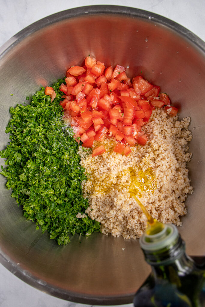 A large mixing bowl filled with the ingredients for tabbouleh not yet mixed while olive oil is being drizzled on to them.