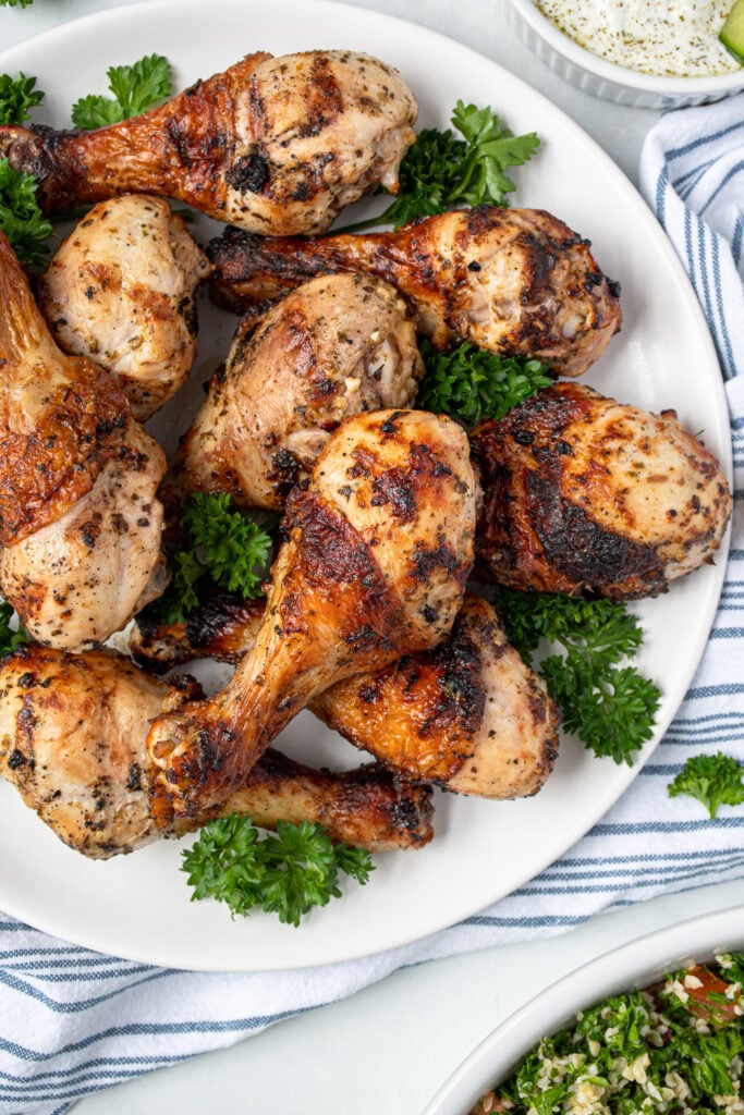 A white plate full of grilled greek marinated chicken drumsticks showing off crispy brown skins.