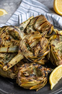 A platter of grilled artichokes with a half of an artichoke on top showing off charred grill marks.