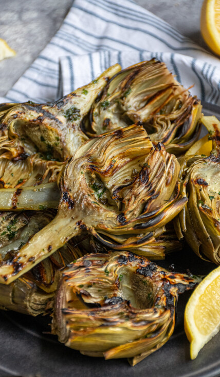 A platter of grilled artichokes with a half of an artichoke on top showing off charred grill marks.