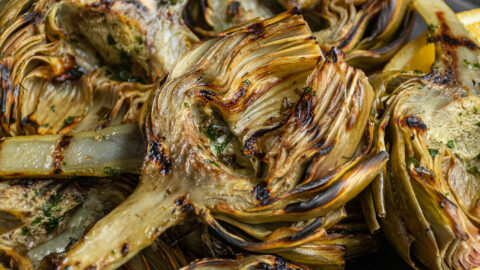 A platter of grilled artichokes with a half of an artichoke on top showing off charred grill marks.