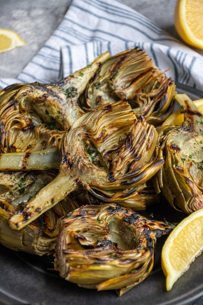 A platter of grilled artichokes with a half of an artichoke on top showing off charred grill marks.
