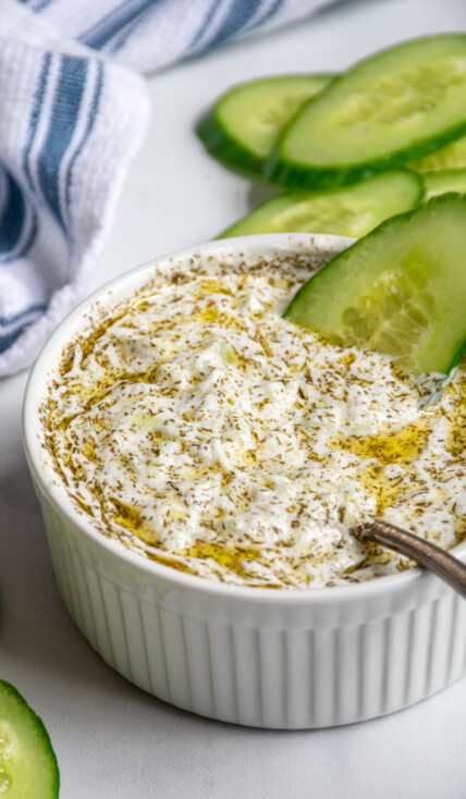 A small white bowl filled with homemade tzatziki sauce dipped with a cucumber slice and a spoon.