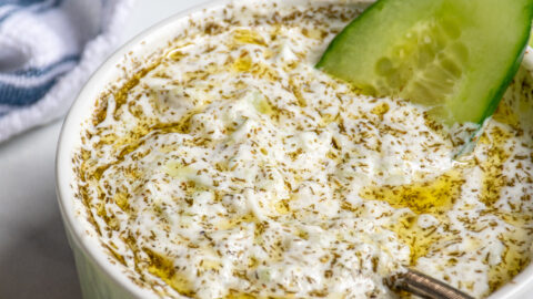 A small white bowl filled with homemade tzatziki sauce dipped with a cucumber slice and a spoon.