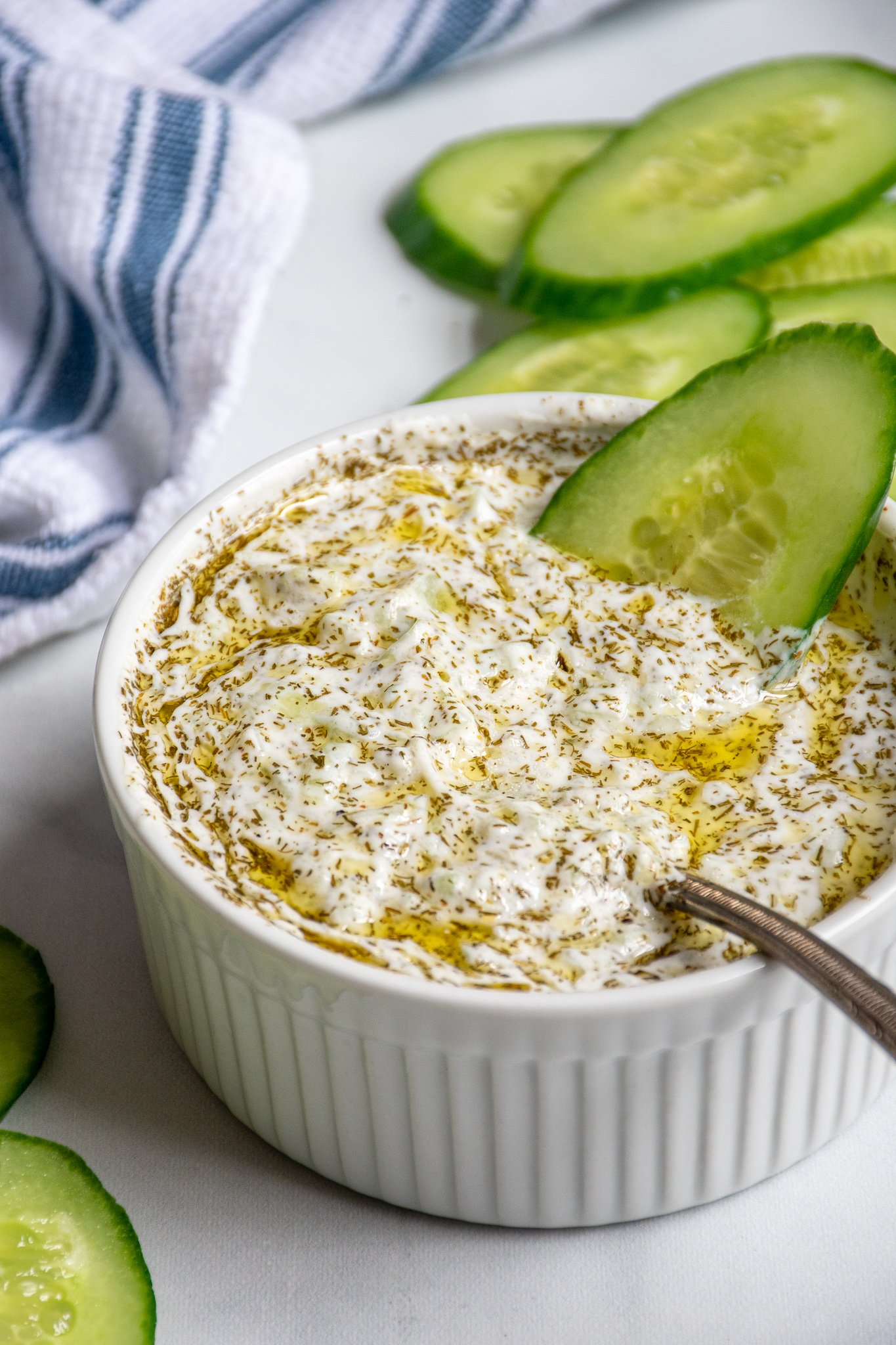 A small white bowl filled with homemade tzatziki sauce dipped with a cucumber slice and a spoon.