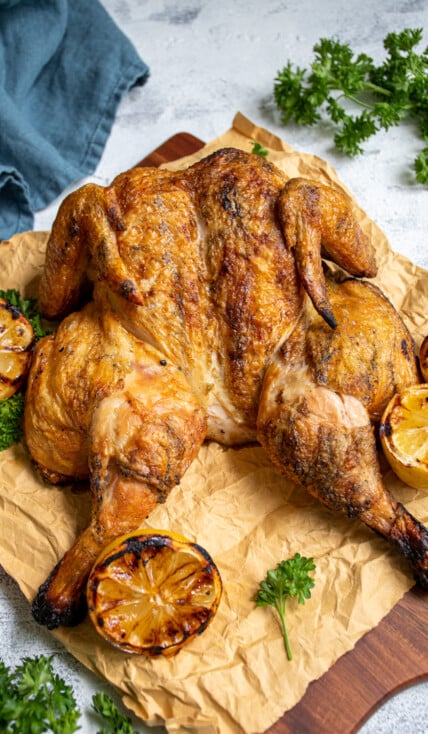 Grilled golden brown spatchcocked chicken sitting on butcher paper and a cutting board next to grilled lemons and parsley.