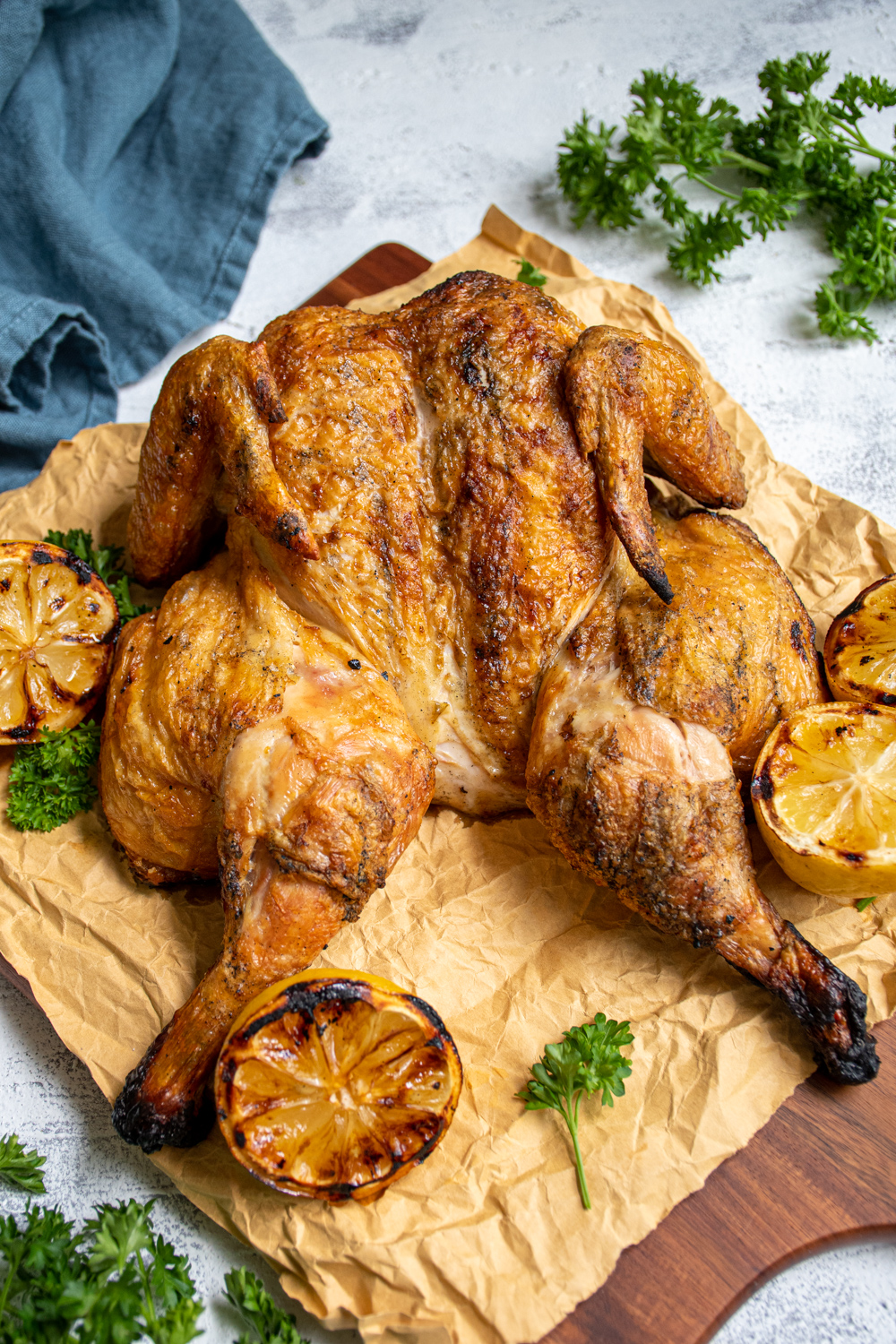 Grilled golden brown spatchcocked chicken sitting on butcher paper and a cutting board next to grilled lemons and parsley.