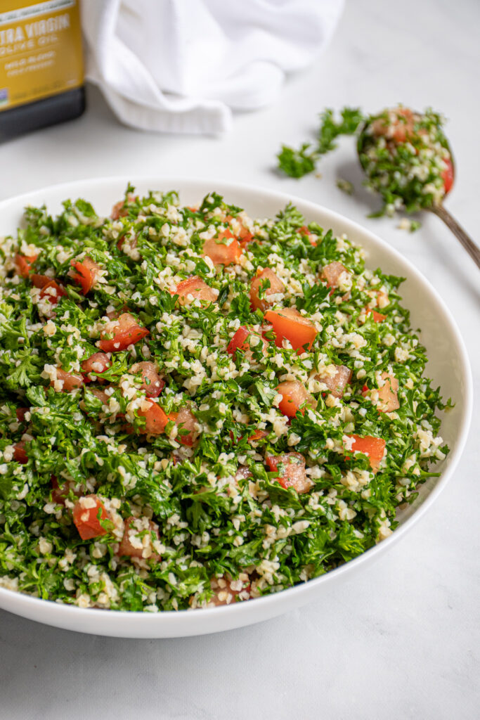 A large white bowl filled with tabbouleh salad with a spoonful out on the side with parsley leaves falling off.