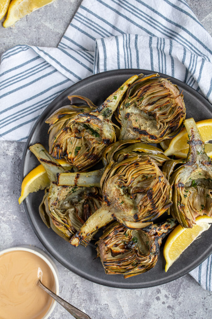 Overhead view of a platter of grilled artichokes fresh from the grill surrounded by cut up lemons and balsamic dipping sauce.
