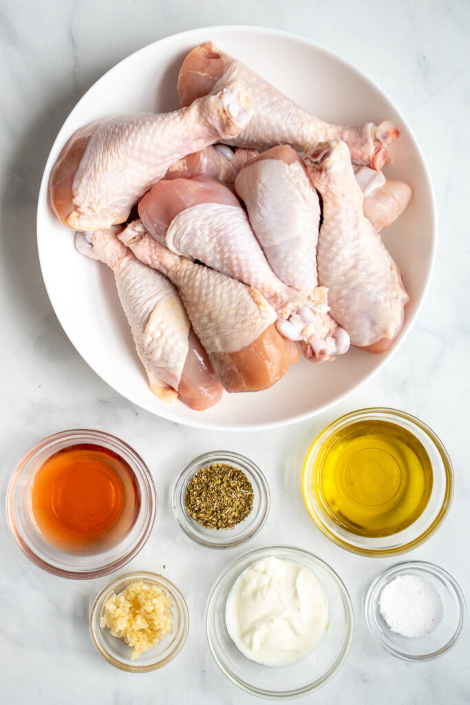 Ingredients needed to make greek marinated chicken laid out on a tabletop with chicken drumsticks.