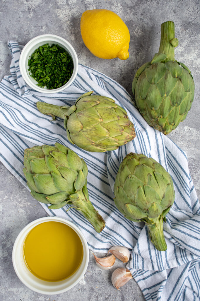 Ingredients needed to grill artichokes including fresh artichokes, lemon, olive oil.