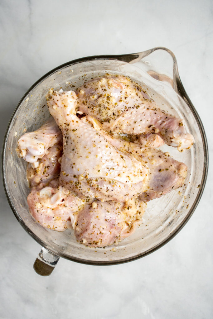 Chicken drumsticks in a large glass bowl covered in greek marinade.